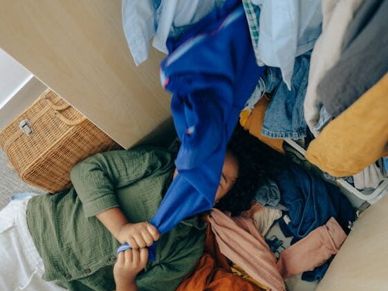 a woman feel exhausted of clutter and lay under pile of clothes overflowed from closet