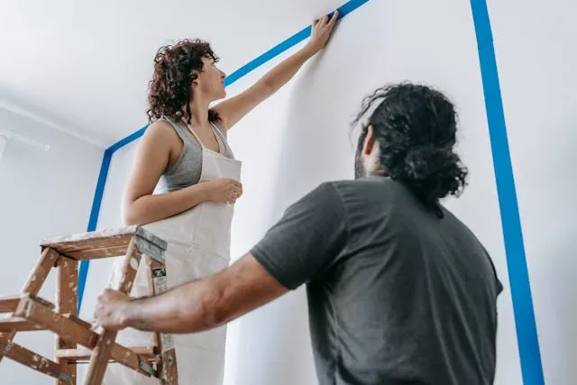 a couple applying painting tape to the edge of the wall